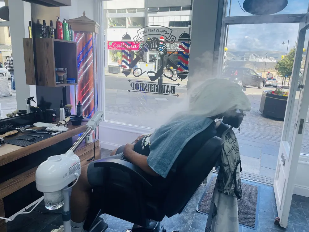 A client reclined in the barber chair under a hot towel with steam rising, beside the shop window
